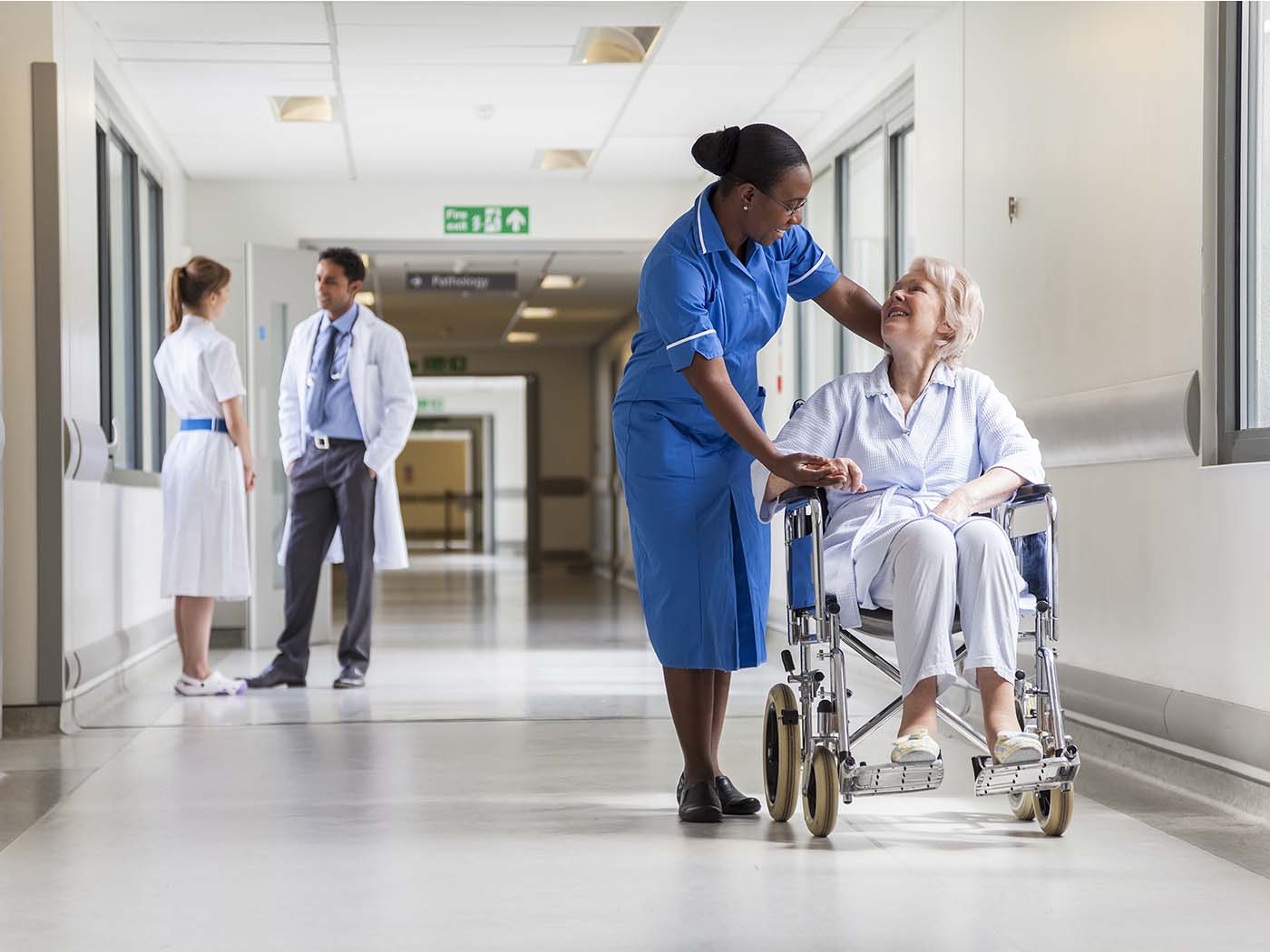 Man sieht eine Frau in einem Rollstuhl, neben ihr eine Krankenschwester. Dahinter stehen ein Arzt und eine Angehörige und unterhalten sich.