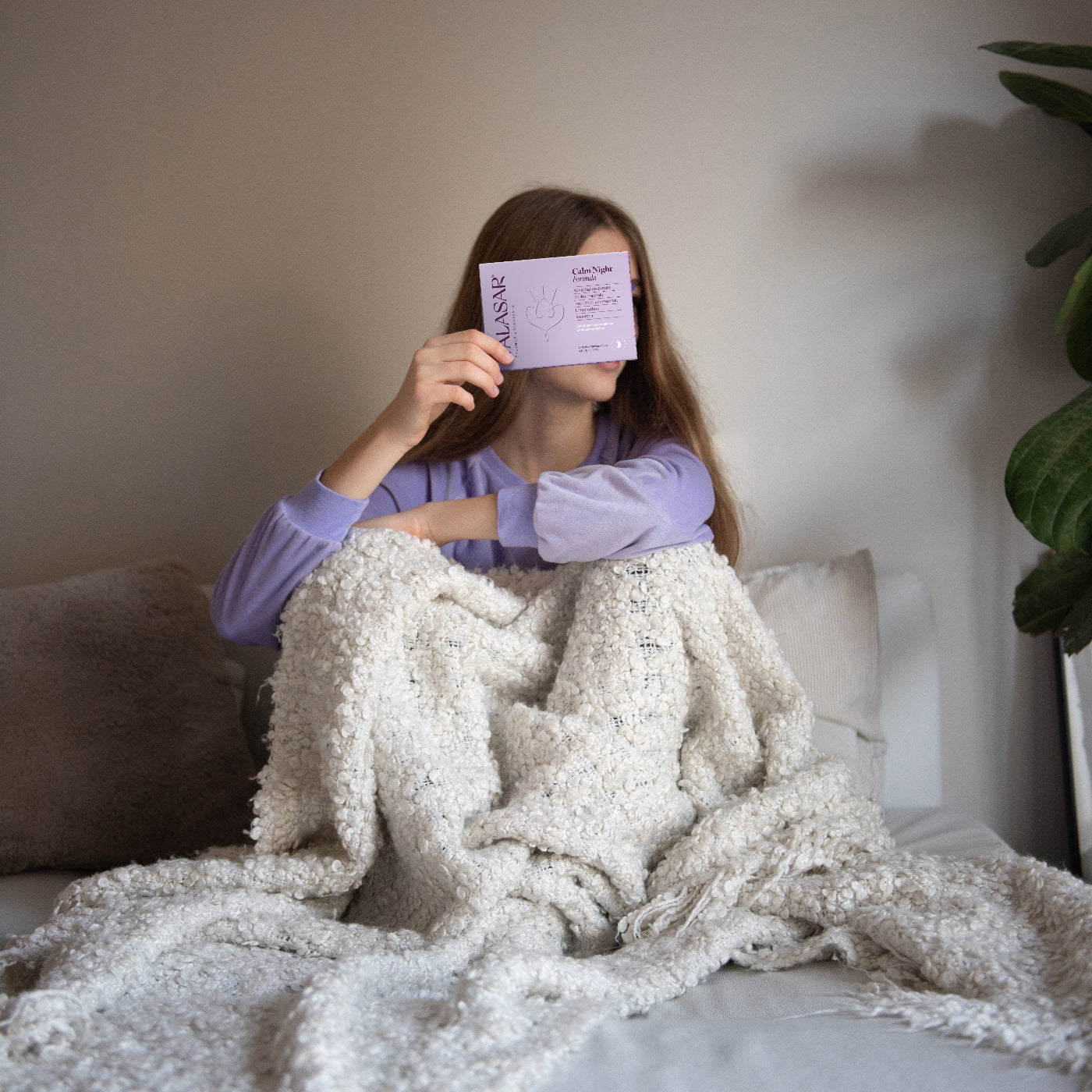 Eine Frau sitzt auf dem Sofa, eingekuschelt in eine Decke und hält das Produkt von Talasar Calm Night hoch.