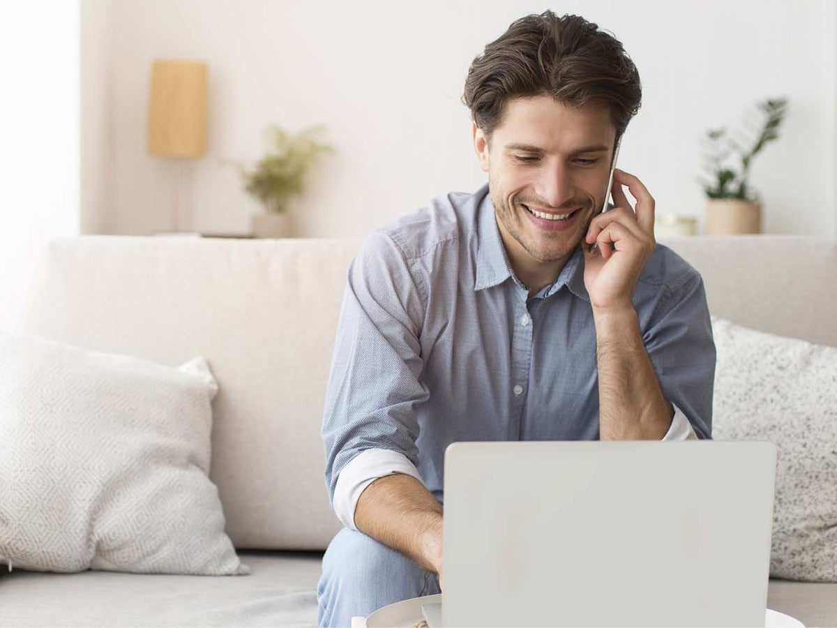 Ein Mann sitzt auf dem Sofa, vor ihm steht ein aufgeklapptes Laptop. Nebenbei telefoniert er.
