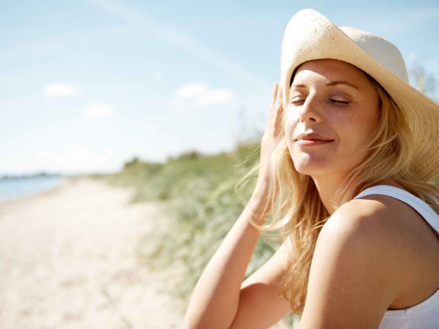 Eine Frau sitzt am Strand. Sie hat die Augen geschlossen und blickt in Richtung der Sonne. Hinter ihr sieht man Dünen.
