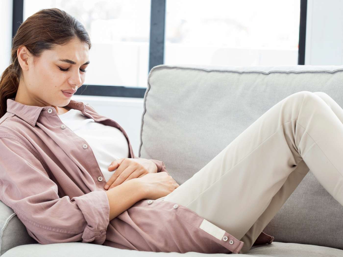Eine Frau liegt auf dem Sofa, die Beine angewinkelt. Sie hält sich den Bauch vor Schmerzen.