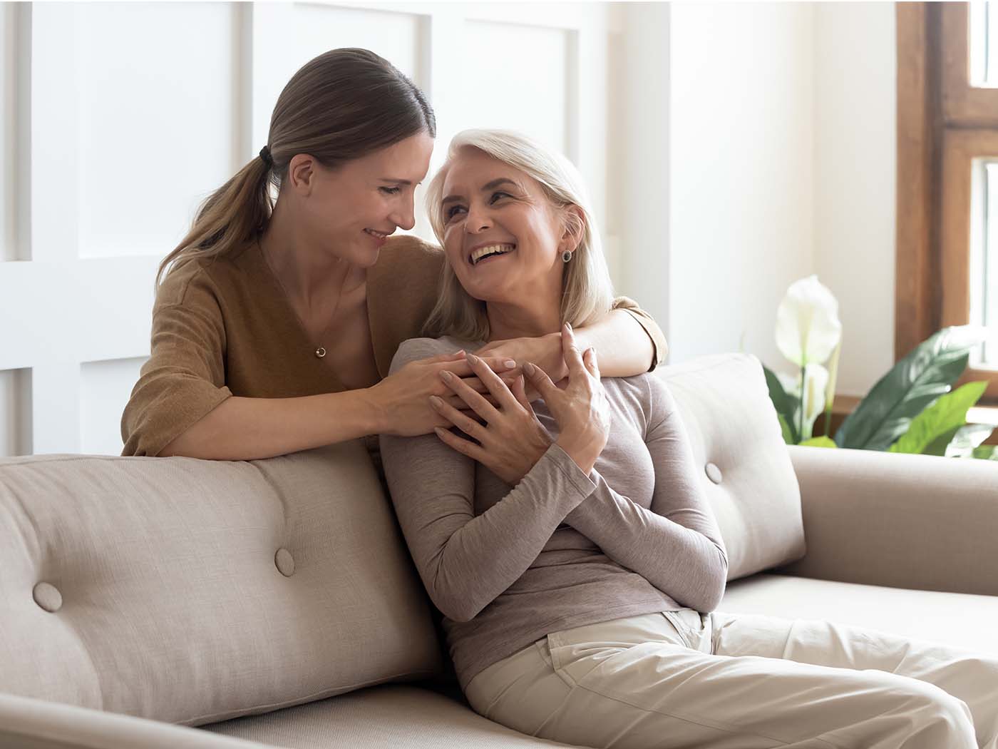 Eine Frau sitzt auf dem Sofa, die andere ist hinter ihr und umarmt sie. Beide lachen.