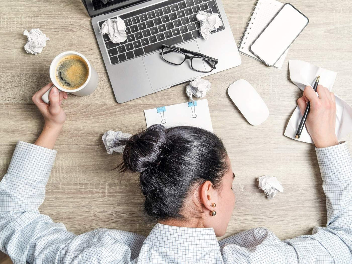 Eine Frau liegt mit dem Oberkörper auf dem Tisch. In der linken Hand hält sie den Henkel der Kaffeetasse, in der rechten einen Stift. Vor ihr steht ihr Computer auf dem ihre Brille liegt. Neben und vor ihr zerknülltes Papier.