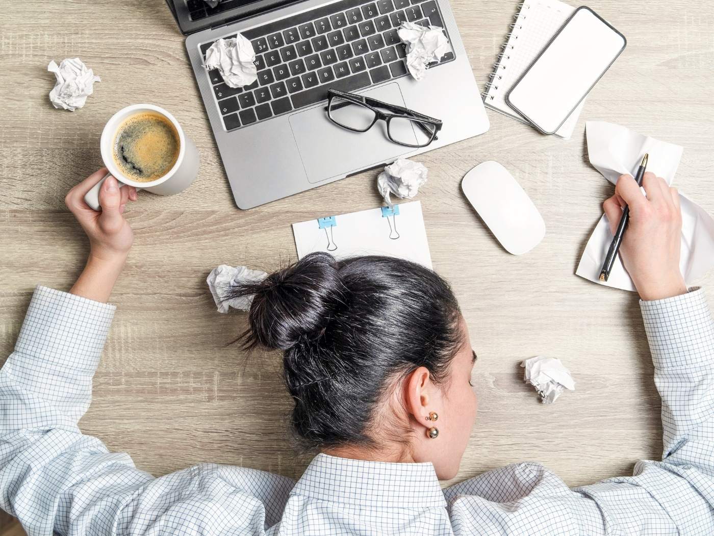 Eine Frau liegt mit dem Oberkörper auf dem Tisch. In der linken Hand hält sie den Henkel der Kaffeetasse, in der rechten einen Stift. Vor ihr steht ihr Computer auf dem ihre Brille liegt. Neben und vor ihr zerknülltes Papier.