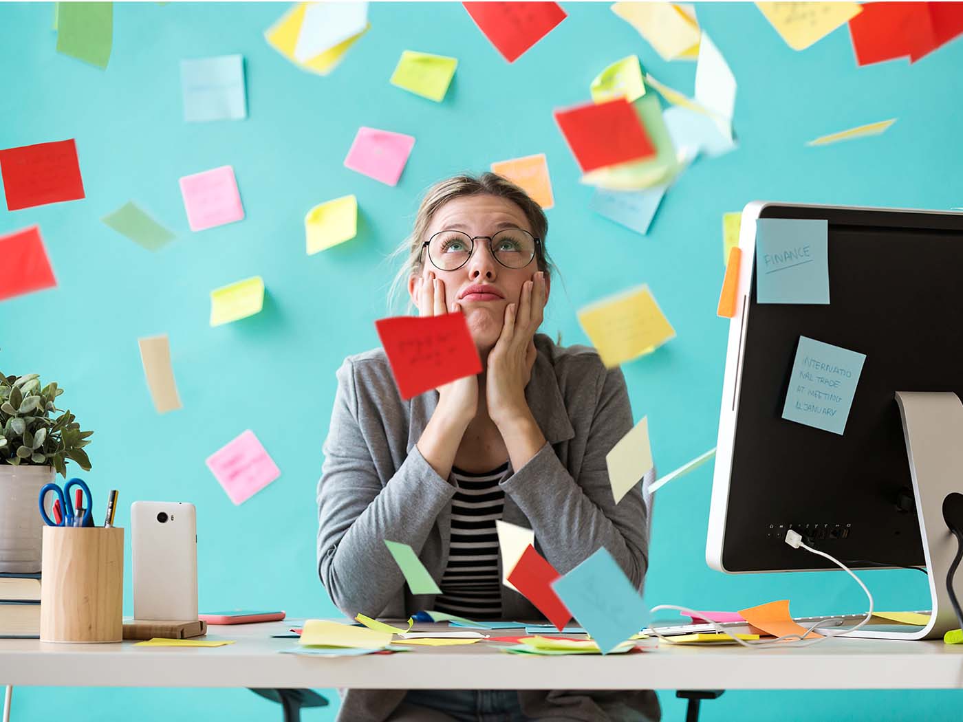 eine Frau sitzt an ihrem Schreibtisch. Mit ihren Händen umfasst sie ihr Kinn. Überall sind Post it's und sie sieht hilfesuchend nach oben.